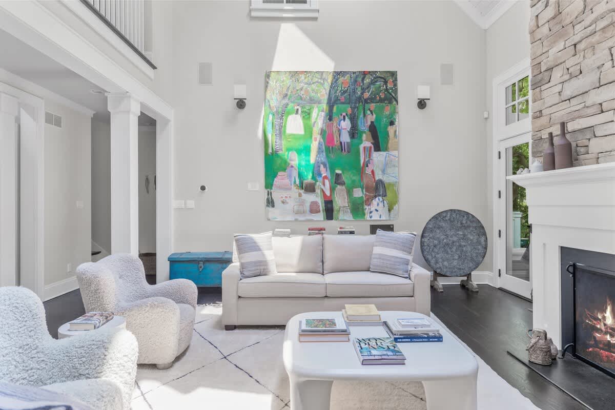 A bright living room with a large abstract painting on the wall above a light-colored sofa. Two fluffy armchairs and a white coffee table are arranged on a patterned rug, with a vintage blue chest adding a pop of color.