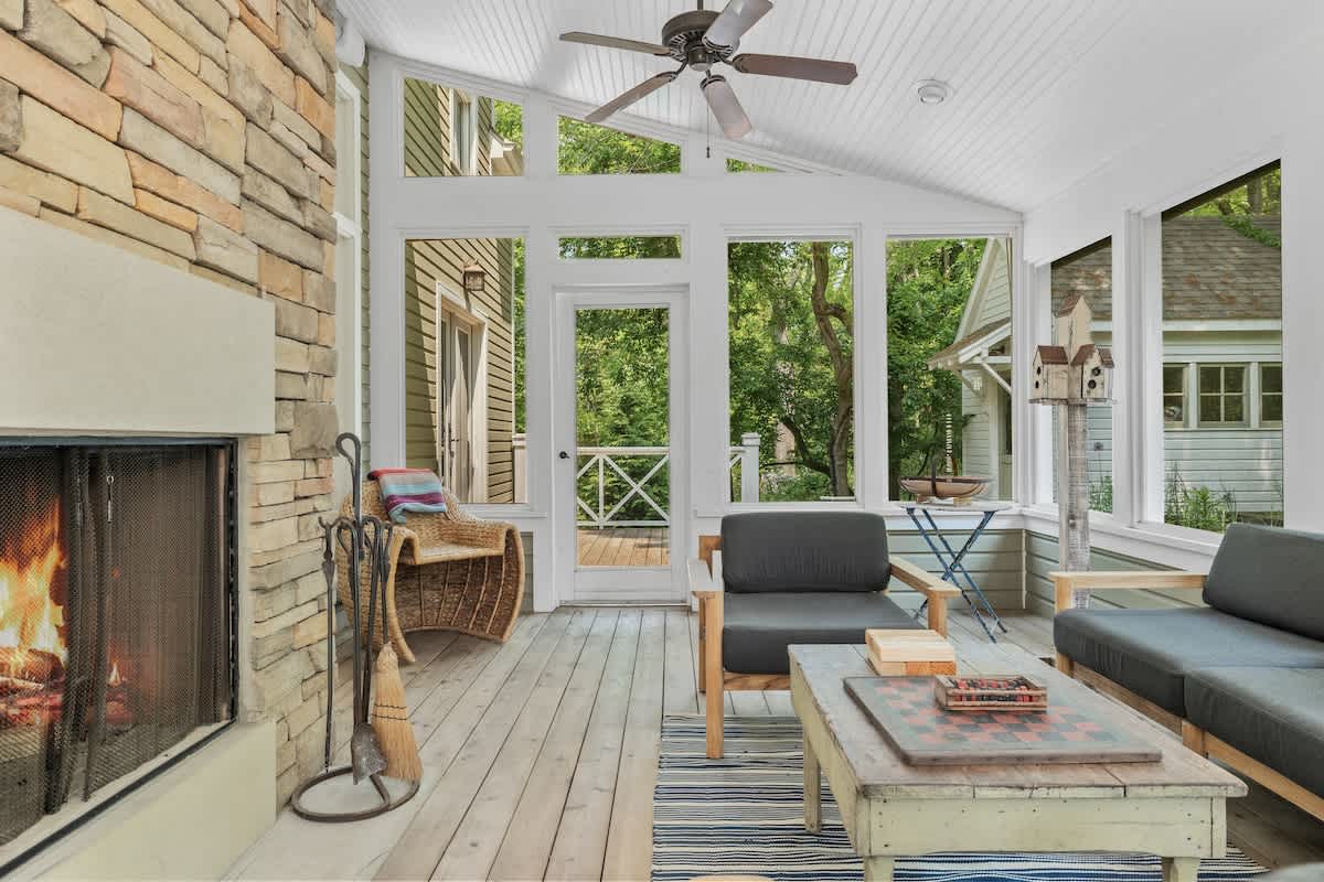 Spacious screened porch with a stone fireplace, a seating area with a coffee table and armchairs, and a door leading to an outdoor deck, all surrounded by lush greenery.