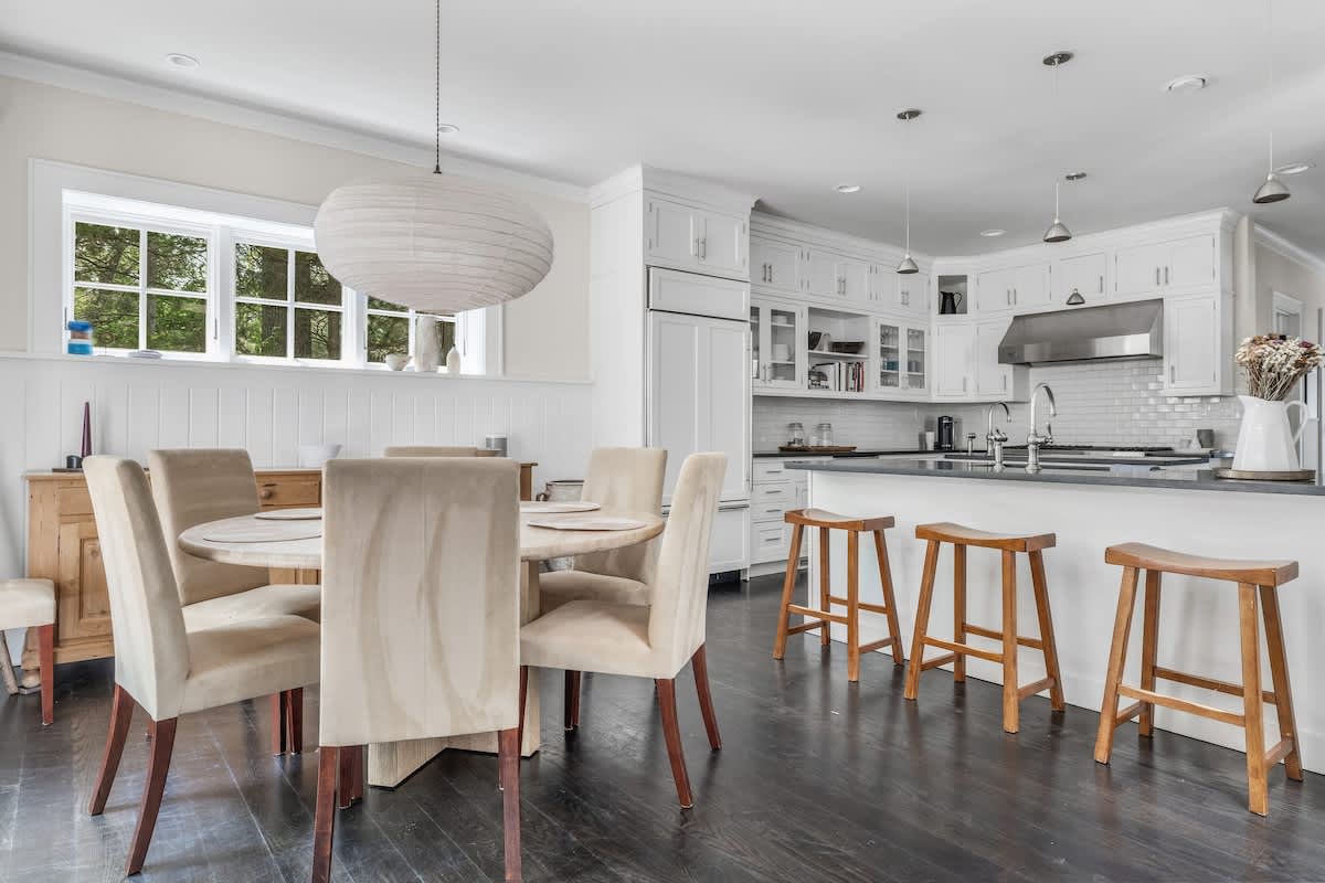 Open-concept dining area with a round wooden table and six upholstered chairs, under a large paper lantern, flowing into a modern white kitchen with a dark countertop island and wooden bar stools.