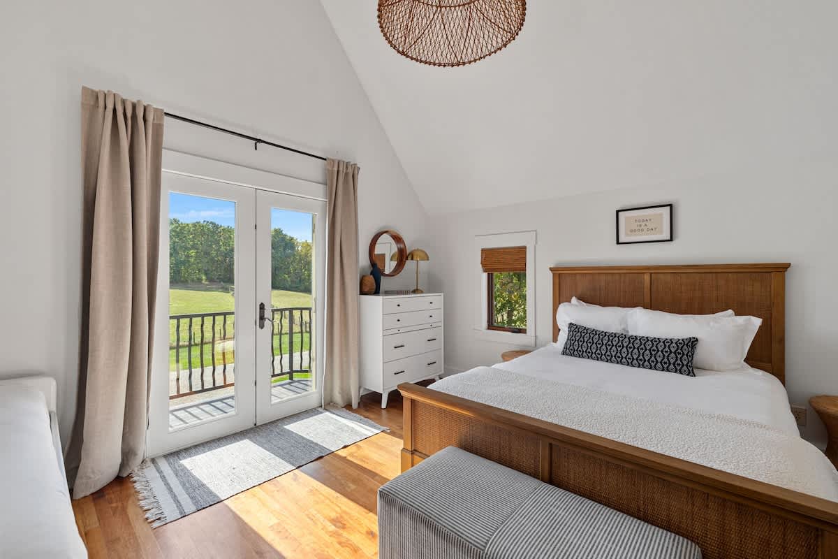 Cozy bedroom featuring a wooden bed frame, a white dresser, and French doors that open to a small balcony with views of the green landscape. A unique woven light fixture hangs from the vaulted ceiling.