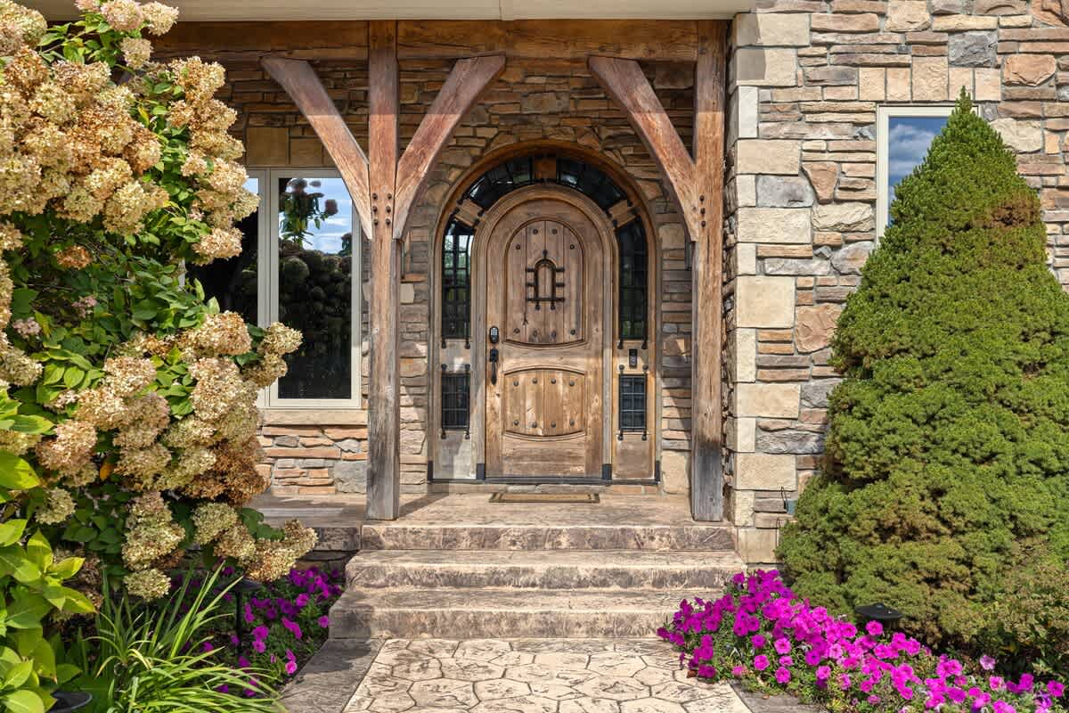 The inviting front entrance showcases a grand arched wooden door with intricate detailing, framed by a rustic stone facade and supported by wooden beams, flanked by vibrant purple flowers and lush greenery.