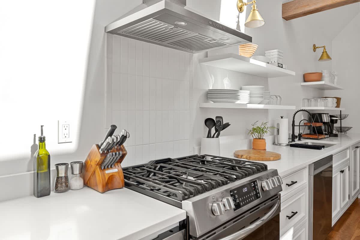 A detailed view of a modern kitchen counter with a gas range, stainless steel hood, white subway tile backsplash, and open shelving displaying dishes, alongside a black faucet and sink.