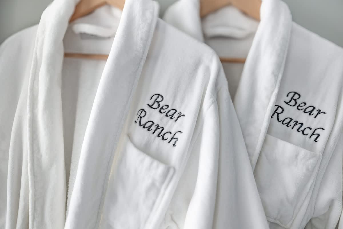 A close-up detail shot of two plush white bathrobes, each embroidered with 'Bear Ranch', hanging neatly on wooden hangers, ready for guest use.