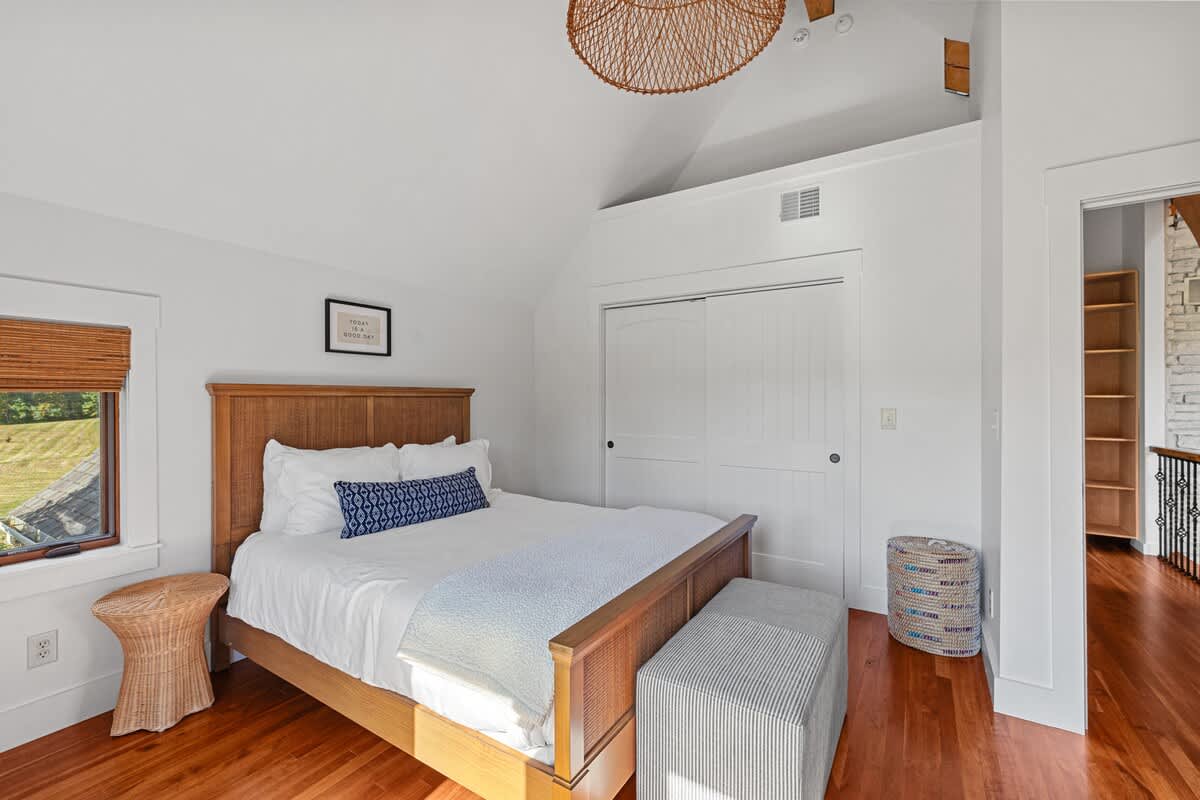 Bright bedroom with white walls, warm hardwood floors, a wooden bed frame, and a striped ottoman at the foot of the bed. A unique woven light fixture hangs from the vaulted ceiling, and a small window offers a view of greenery.