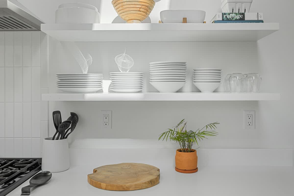 A modern kitchen features white open shelving displaying neatly stacked white dishes and bowls, a wooden cutting board on a white countertop, and a small potted plant adding a touch of greenery.