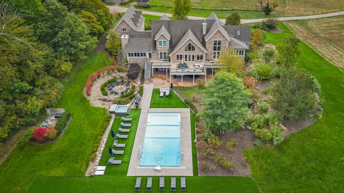 An aerial perspective reveals the rear of the large stone house with a spacious deck, a hot tub, and a rectangular private pool, all set within meticulously manicured green lawns and diverse landscaping.