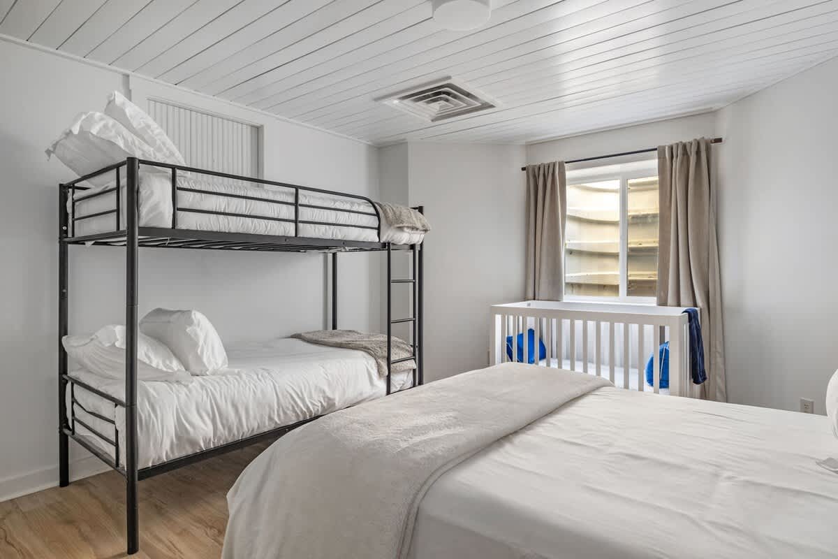 Spacious bedroom with light wood-look flooring and white walls, featuring a black metal bunk bed and a queen bed, both with white bedding. A white crib is placed by a window with a window well.
