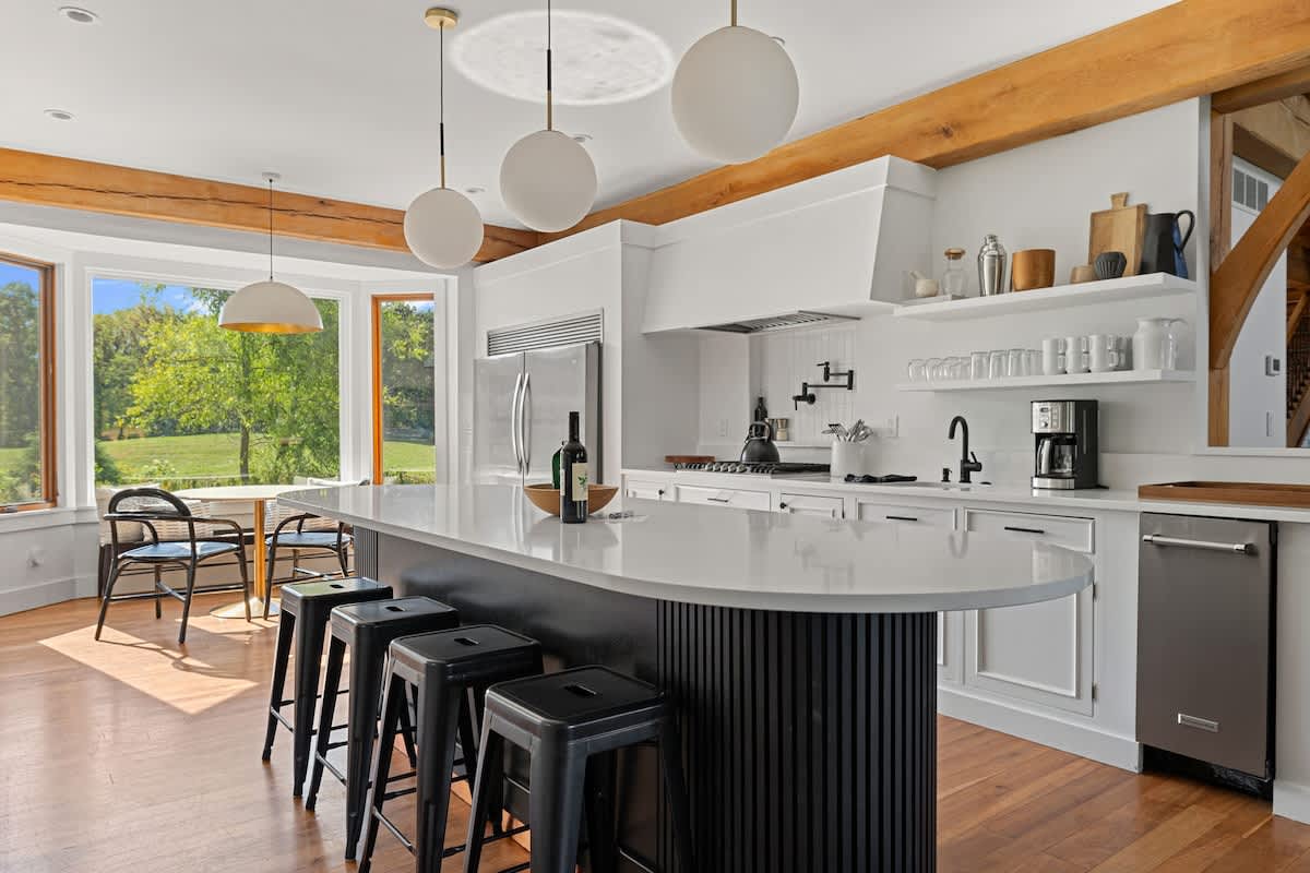 A modern kitchen with a large black island, white countertops, and white cabinetry, featuring three spherical pendant lights, wooden beams, and a bay window with a small breakfast nook.