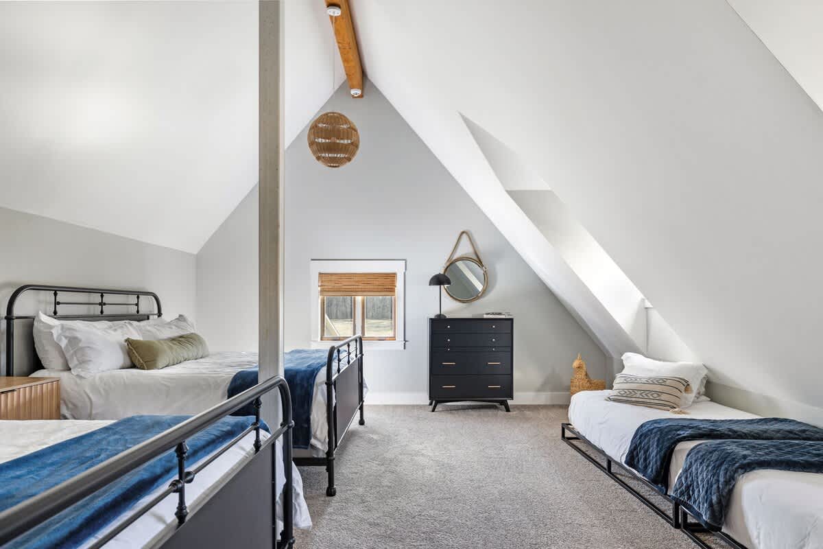 A bright attic bedroom with sloped ceilings and white walls features two black metal-frame beds with white bedding and blue throws, a dark dresser, and a round mirror.