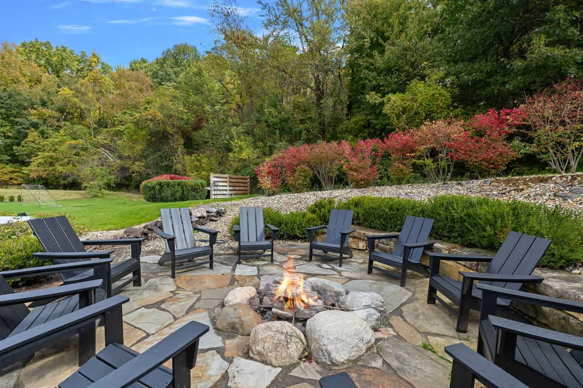 A cozy outdoor fire pit area features a circular flagstone patio with several black Adirondack chairs arranged around a central stone fire pit, nestled within vibrant green and autumnal landscaping.