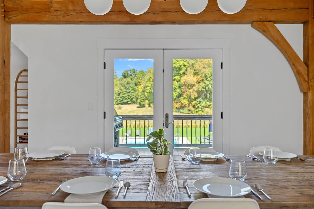 Dining table elegantly set with plates and glasses, offering a picturesque view through French doors to an outdoor deck. A grill and a glimpse of a swimming pool are visible beyond the doors.