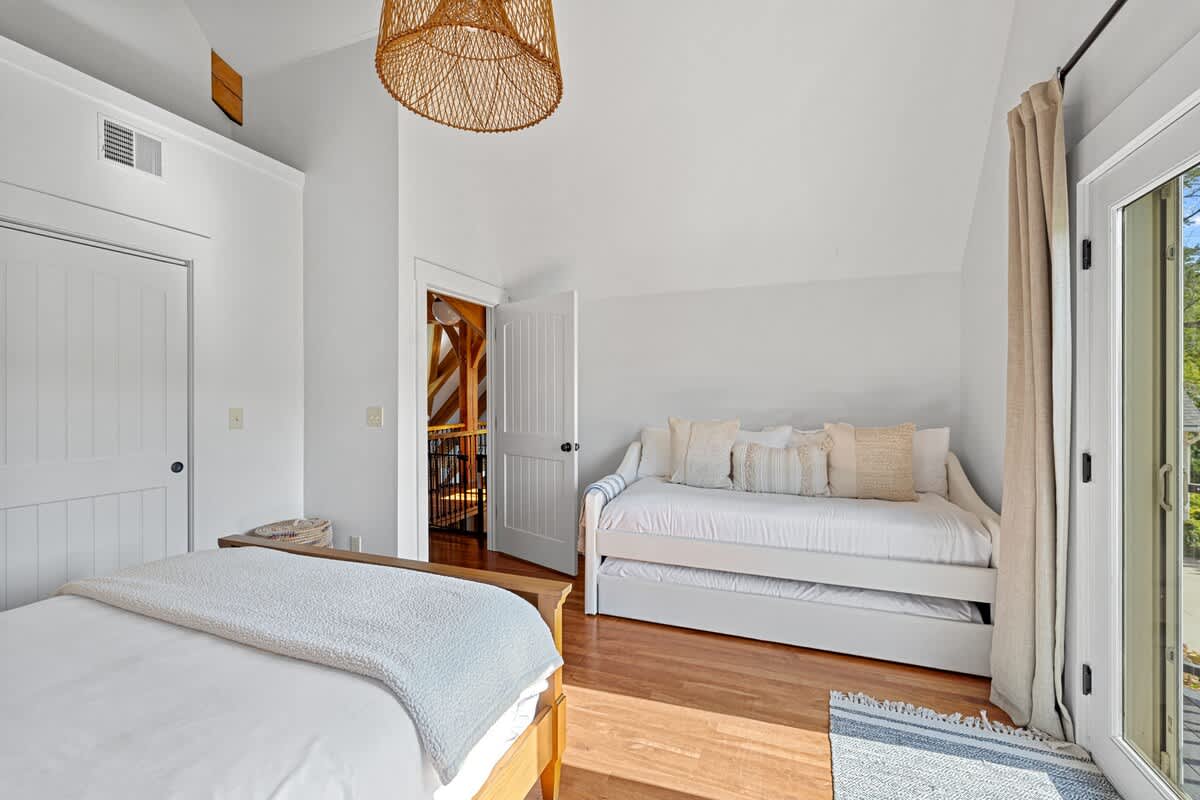 White-walled bedroom featuring a daybed with a trundle, warm hardwood floors, and a door leading to a balcony. A woven light fixture hangs from the vaulted ceiling, and a small patterned rug is placed on the floor.