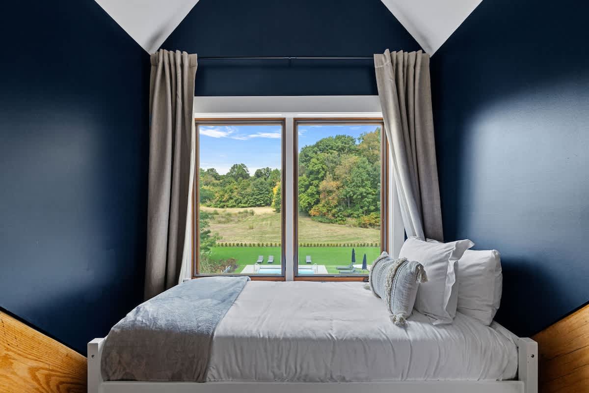 Close-up view of a twin bed in a bedroom with dark blue walls and warm hardwood floors. A large window provides expansive views of the green outdoor landscape, framed by light-colored curtains.