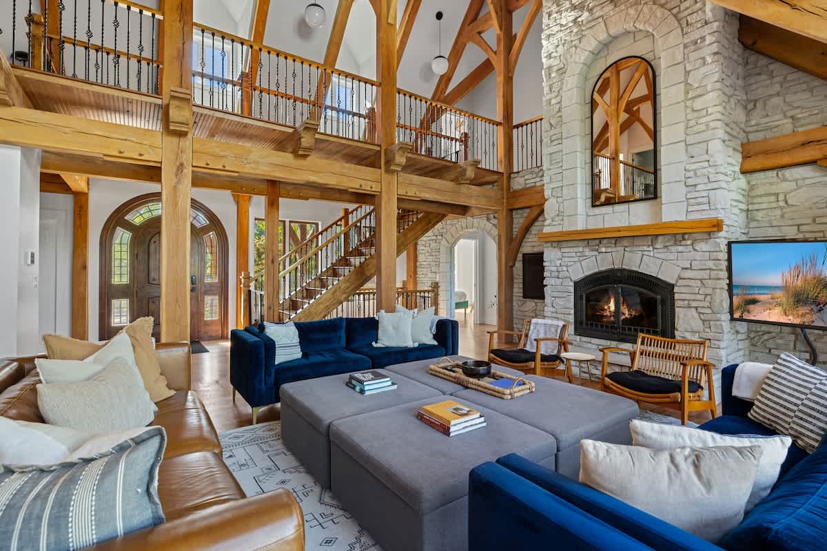 Cozy living room centered around a large grey ottoman, flanked by blue velvet sofas and a brown leather sofa. A grand stone fireplace and exposed timber beams create a rustic yet luxurious ambiance.