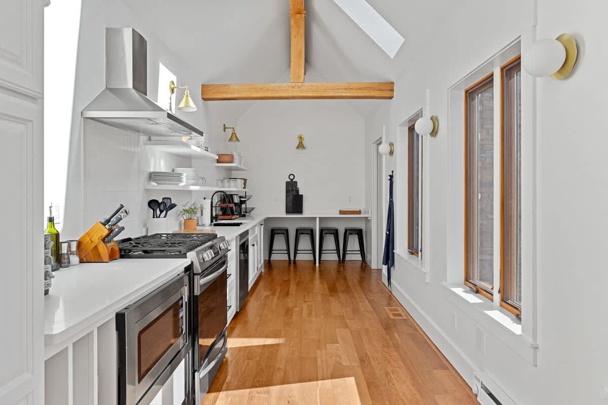 A bright, narrow kitchen or prep area with white walls, vaulted ceilings, and exposed wooden beams, featuring a gas range, stainless steel hood, open shelving, and a counter with bar stools.