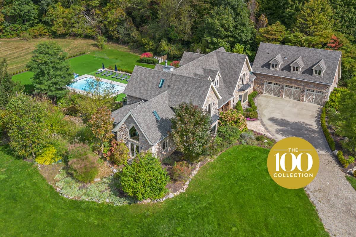 An expansive aerial view showcases a large stone and shingle-roofed luxury home with a detached garage, a rectangular private pool, and lush green lawns surrounded by mature trees and landscaping.