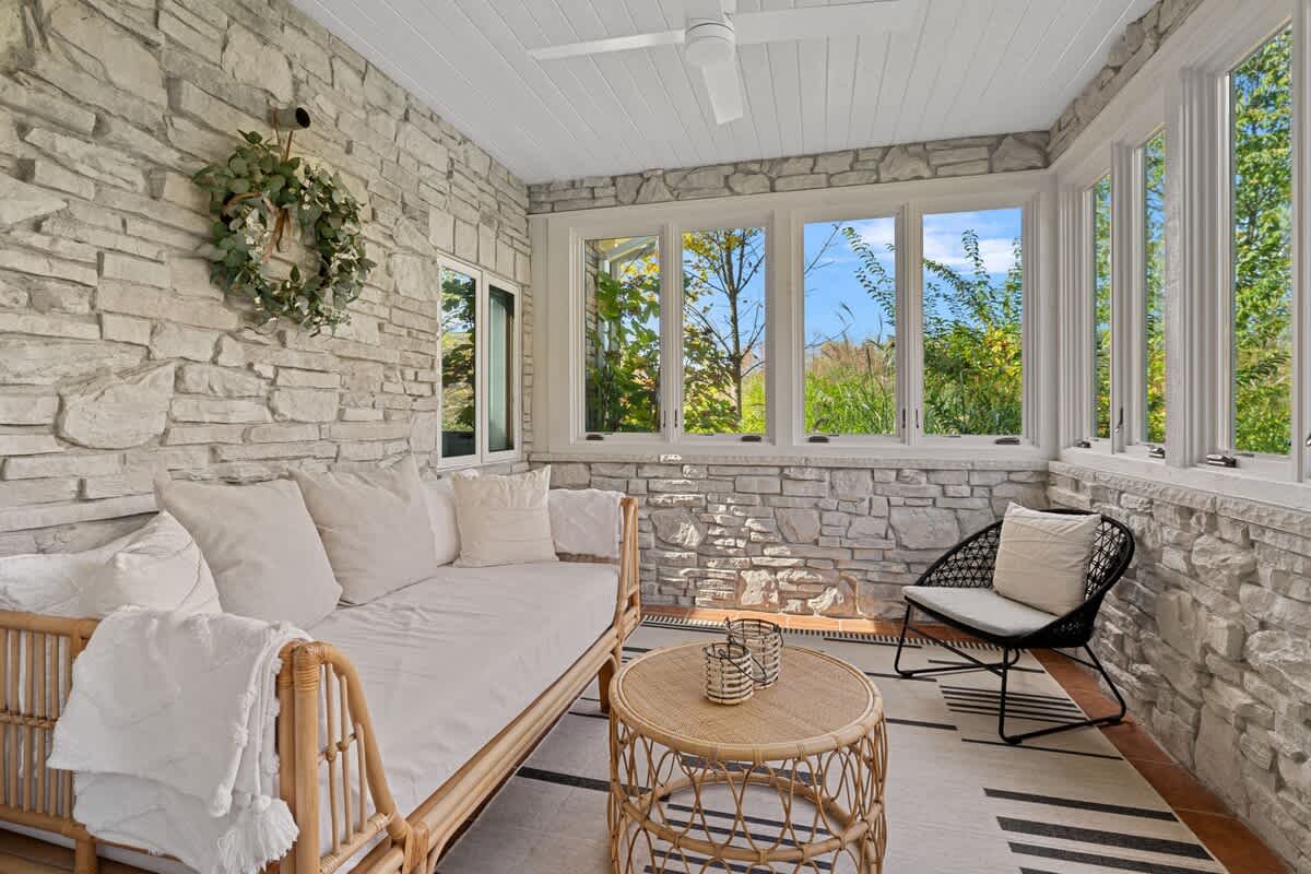 A charming enclosed porch or sunroom features textured stone walls, a white slatted ceiling with a fan, a rattan daybed with white cushions, and large windows offering bright views of the surrounding greenery.