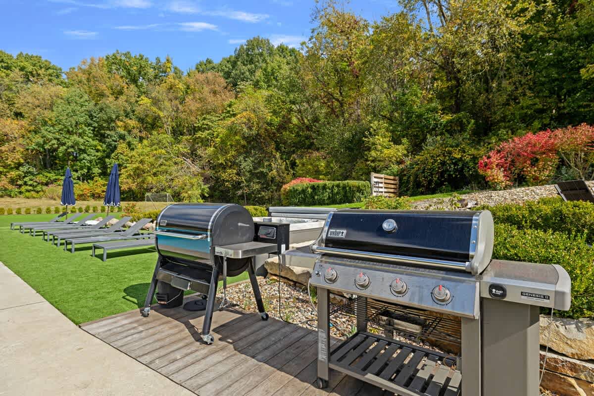 An outdoor grilling station features two large BBQ grills on a wooden deck, adjacent to a manicured lawn with several lounge chairs and umbrellas, surrounded by dense trees.