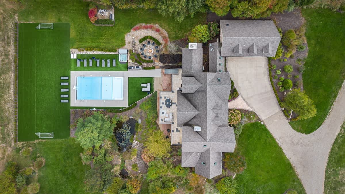 A top-down aerial view reveals the sprawling property, highlighting the main house, a detached garage, a rectangular outdoor pool, a hot tub, and a fire pit area, all surrounded by artificial turf and natural landscaping.