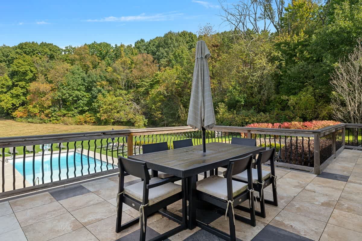 Outdoor deck with patterned tile flooring, featuring a dark wooden dining table with six chairs and an umbrella. The deck overlooks a swimming pool and a vast, tree-lined green landscape under a clear sky.