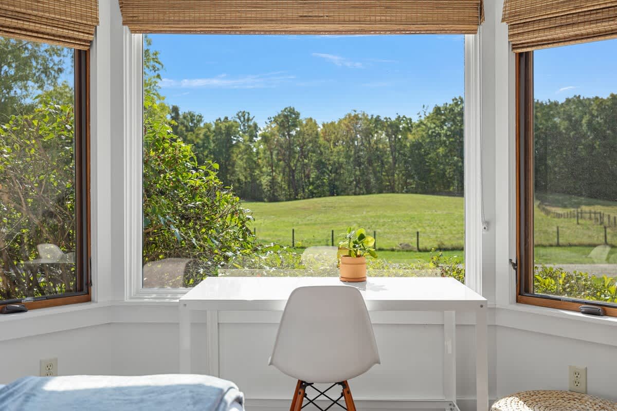 Close-up of a bay window in a bedroom, featuring woven blinds, a white desk, chair, and a potted plant, overlooking a lush green field and distant trees under a clear blue sky.