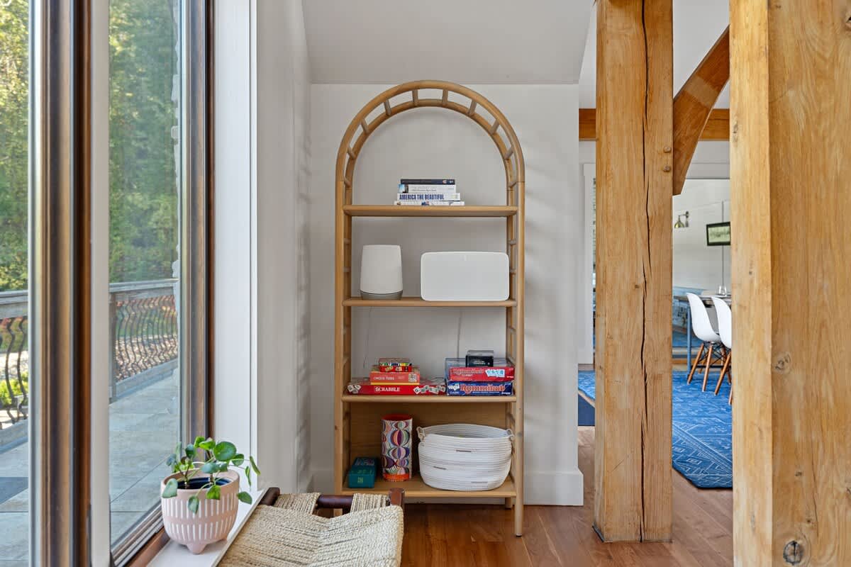 A tall, arched wooden bookshelf filled with books and board games stands beside a large window, offering a glimpse of the outdoor deck. A small potted plant adds a touch of greenery to the windowsill.