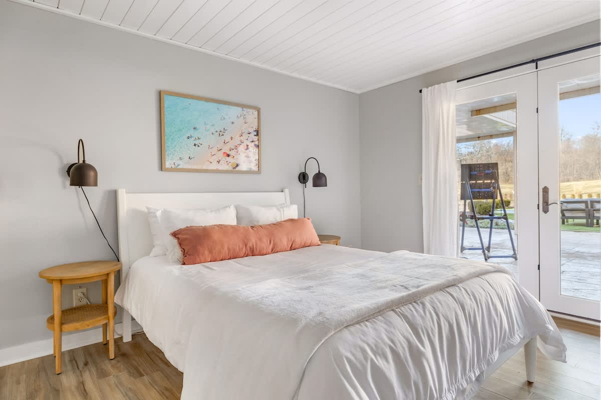 Serene bedroom with light wood-look flooring and light gray walls, featuring a white queen bed with a coral bolster pillow and two wall-mounted sconces. French doors open to an outdoor patio area.