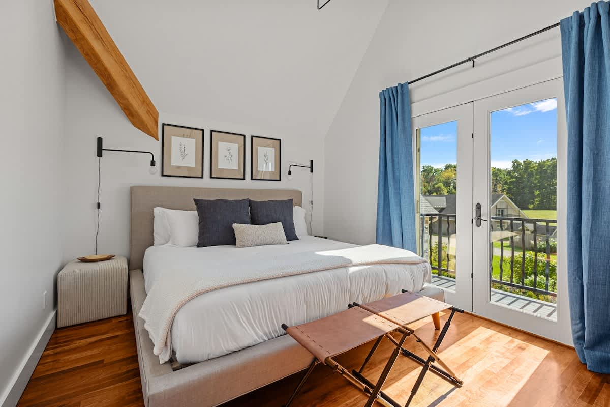 Elegant bedroom with a king-sized bed, light gray upholstered headboard, and rich hardwood floors. French doors open to a balcony with views of the surrounding landscape, framed by long blue curtains.