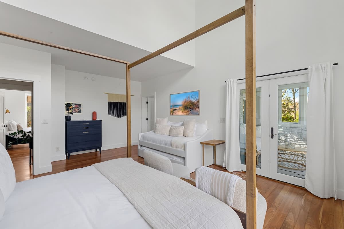 A spacious bedroom with a prominent four-poster bed, white bedding, and a daybed with a mounted television. French doors open to an outdoor area, complemented by light hardwood floors and a vaulted ceiling.