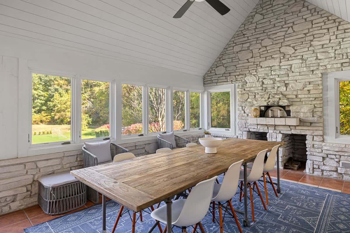 A bright dining room with a long rustic wooden table and modern chairs, surrounded by large windows offering views of lush greenery, and featuring a stone fireplace with a built-in pizza oven.