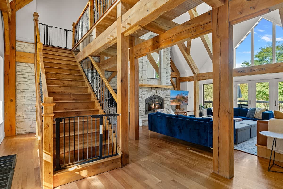 Spacious living area with a grand wooden staircase, exposed timber beams, and a prominent stone fireplace. A blue sofa and a television are visible in the background, with a baby gate at the bottom of the stairs.
