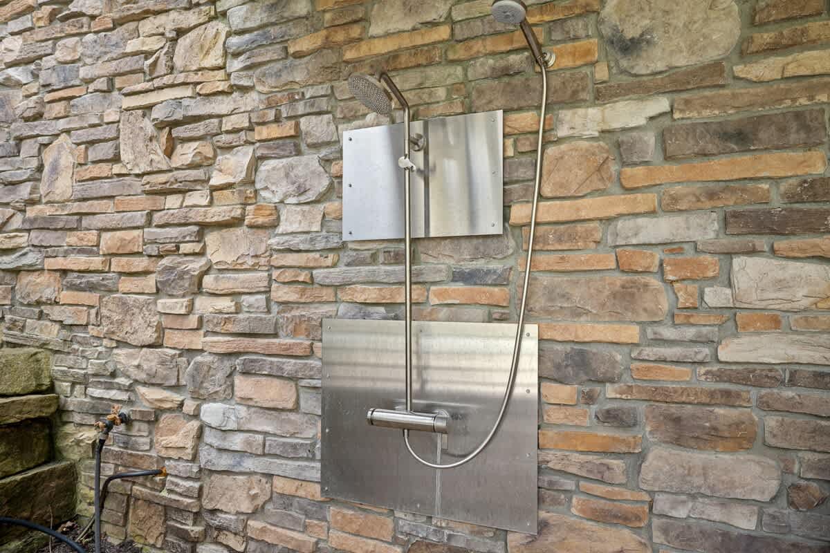 An outdoor shower with stainless steel fixtures is mounted on a rustic stacked stone wall, featuring a handheld showerhead and a mixer.