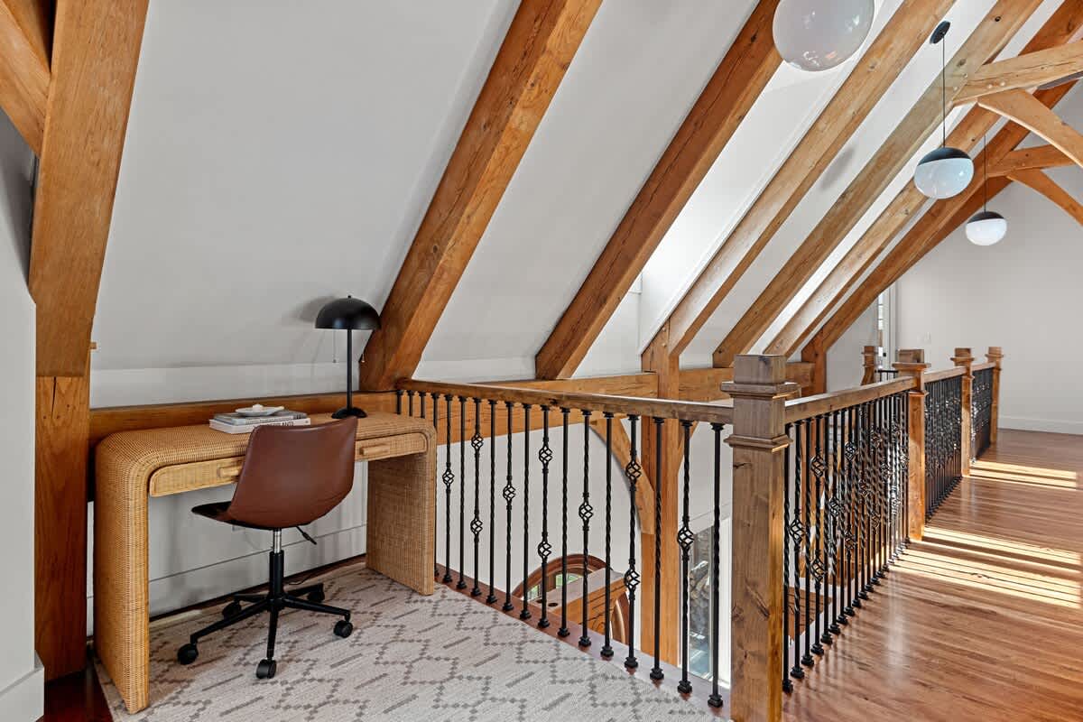 Dedicated loft workspace area with a rattan desk and an ergonomic office chair. It is situated under a sloped ceiling with prominent exposed wooden beams, overlooking a lower level through a decorative railing.
