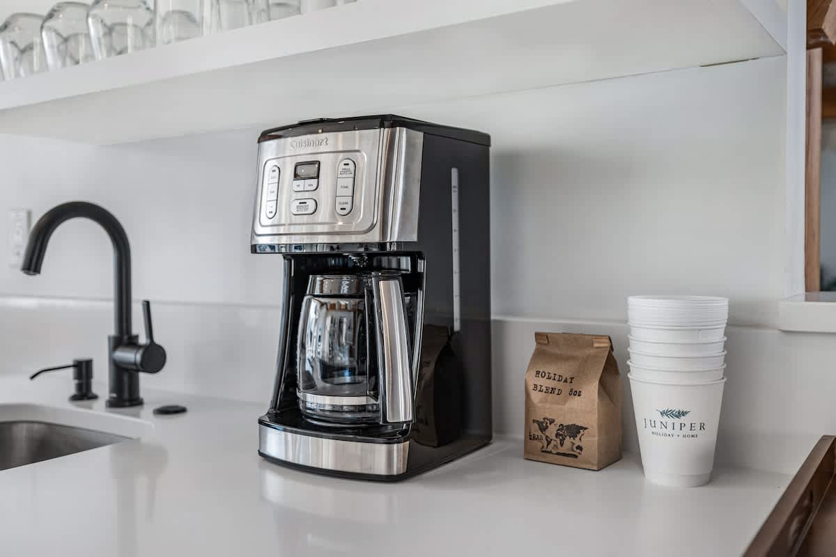 Close-up of a kitchen counter with a Cuisinart coffee maker, a black sink with a gooseneck faucet, and white disposable cups next to a bag of 'Holiday Blend' coffee.