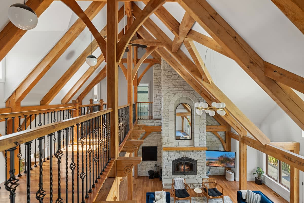 A grand living space features a soaring vaulted ceiling with intricate exposed wooden beams, a massive stone fireplace as the focal point, and a mezzanine balcony with decorative wrought iron railings overlooking the room.