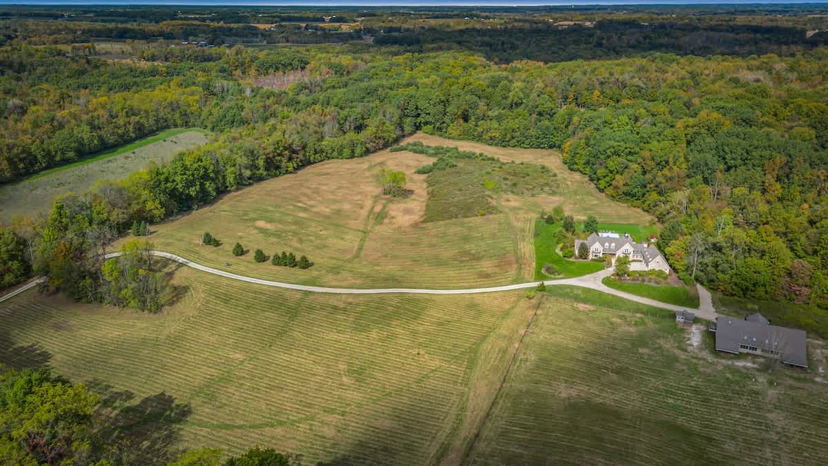 A wide aerial shot displays the vast estate, featuring the main house and its surrounding manicured lawns, a long winding driveway, and extensive natural woodlands and fields in the distance.