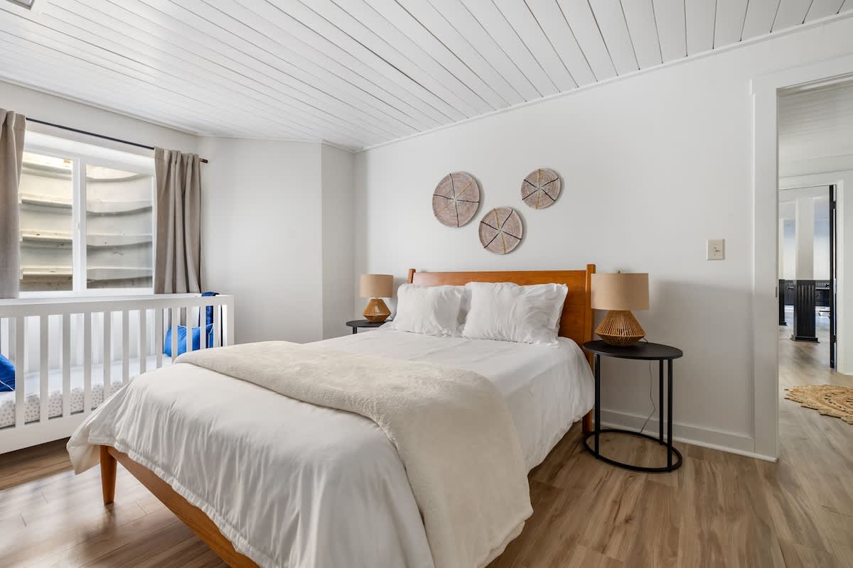 Bright bedroom with light wood-look flooring and white walls, featuring a wooden queen bed with white bedding and two round side tables with lamps. A white crib is positioned by a window with a window well.
