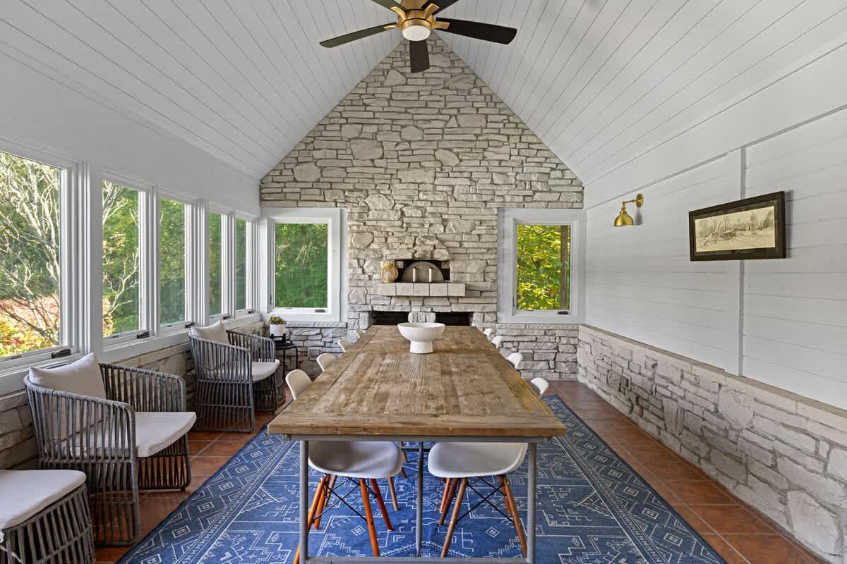 Enclosed dining area or sunroom featuring rustic stone walls, a vaulted white ceiling, and large windows offering outdoor views. A long wooden table is surrounded by chairs, with a built-in pizza oven as a focal point.