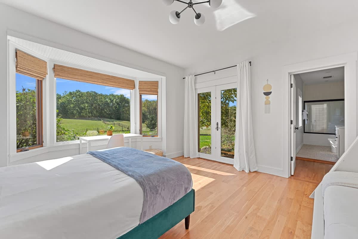 Bright bedroom with light hardwood floors and a large bay window offering expansive green field views. A white desk and chair are placed in the bay window, and a glass-paned door opens to the outdoors.