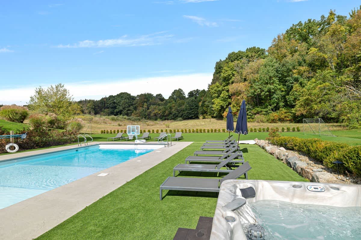 A rectangular outdoor swimming pool with concrete surround is flanked by artificial turf, several dark lounge chairs, and a bubbling hot tub in the foreground. A soccer goal is visible in the distance.