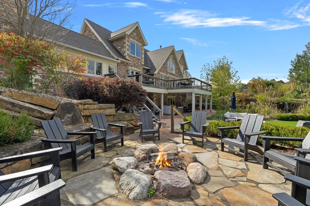An inviting outdoor fire pit area with a flagstone patio is surrounded by several dark Adirondack chairs, set against a backdrop of a large house and lush, terraced landscaping.