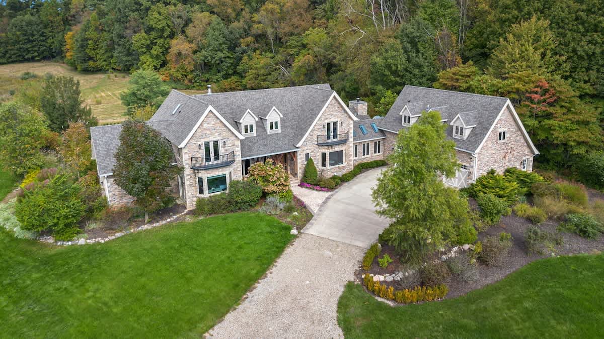 An aerial view showcases the large stone and siding house with multiple gables and dormer windows, situated amidst extensive green lawns and mature trees. A long gravel driveway leads to the property.