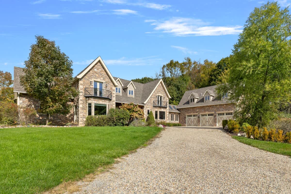 The grand stone and siding facade of the house is visible from a gravel driveway, featuring multiple gables, dark-framed windows, and a detached garage with three bays. Lush landscaping surrounds the entrance.