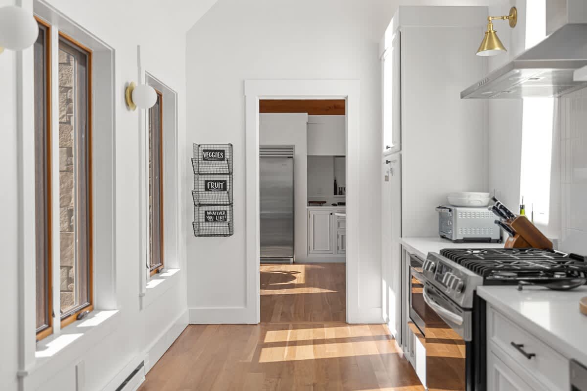 A bright, narrow kitchen hallway with light hardwood floors and white walls, leading to a stainless steel refrigerator visible through a doorway. Wall-mounted wire baskets provide storage on the left.