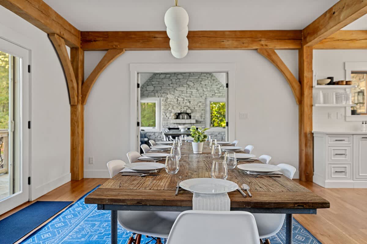 Rustic dining room featuring a long wooden table set for multiple guests with modern white chairs and a contemporary chandelier. Exposed timber beams and a vibrant blue patterned rug define the space.