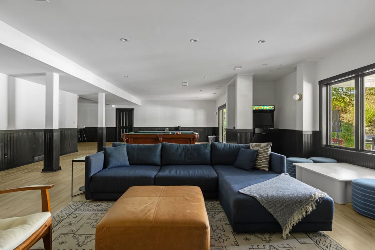 Spacious lower-level living area featuring a large navy sectional sofa and a brown leather ottoman on a patterned rug, with the game room visible in the background. The space has light wood-look flooring and black wainscoting.