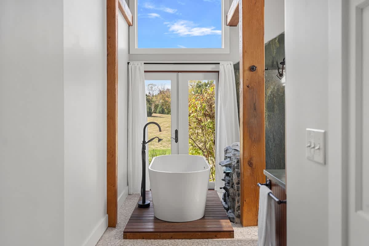 A bright and airy bathroom featuring a freestanding white bathtub on a wooden slatted platform, positioned in front of French doors that open to a serene outdoor landscape. Exposed wooden beams frame the space.