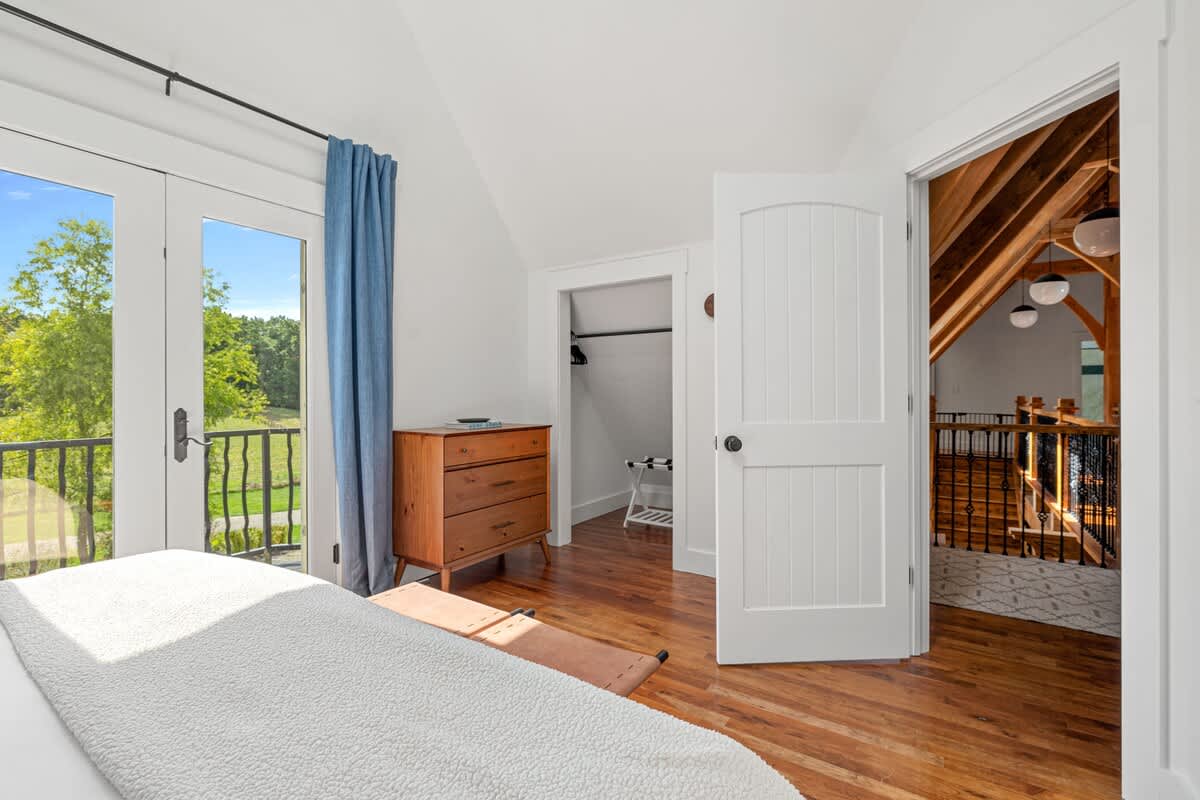 View of a bedroom with warm hardwood floors, a rustic wooden dresser, and an open closet area. French doors lead to a balcony with a scenic outdoor view, complemented by blue curtains and a vaulted ceiling.