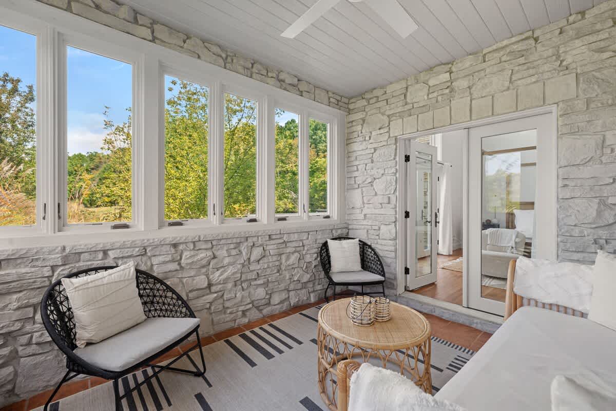 A bright enclosed porch or sunroom with textured stone walls and large windows offering views of lush greenery. The space is furnished with two black woven chairs and a round rattan coffee table.