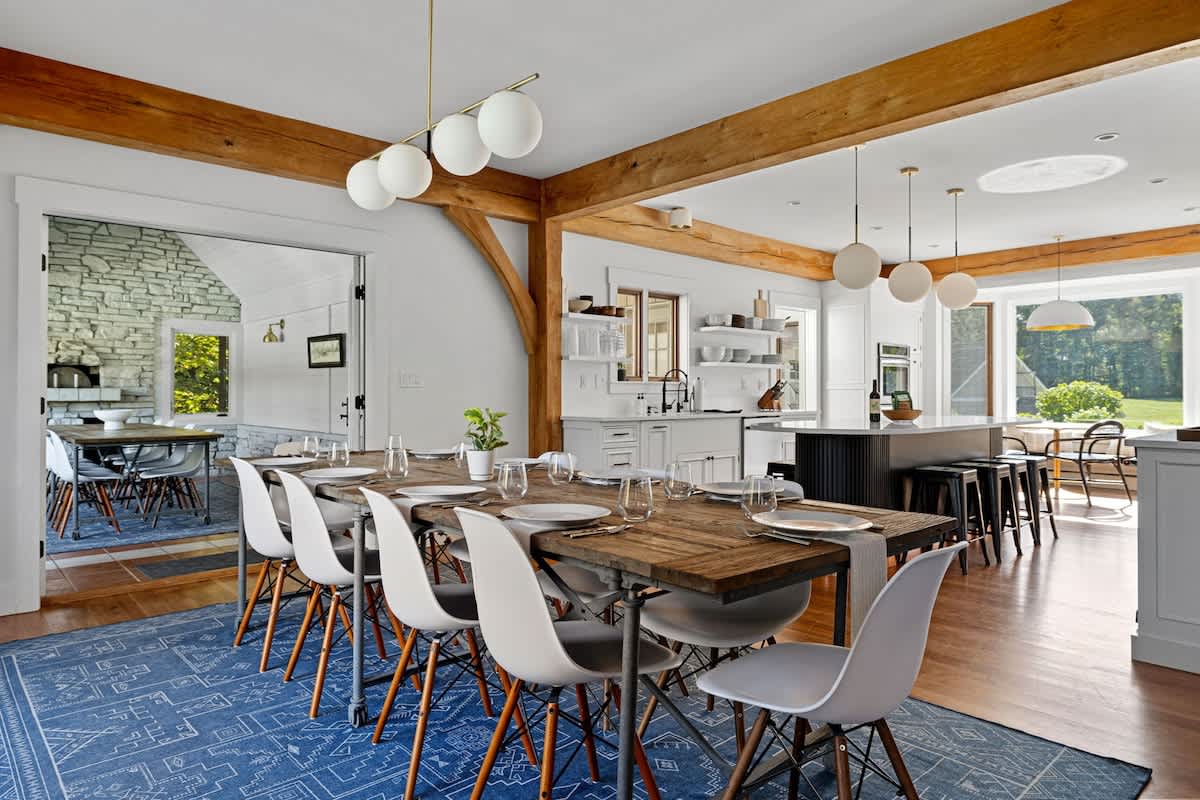 An open-concept dining area features a long rustic wooden table with modern white chairs, set on a blue patterned rug, flowing into a contemporary kitchen with white cabinetry, a large island, and exposed wooden beams overhead.